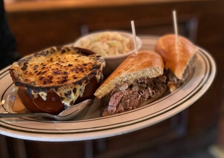 A small bowl of French onion soup next to a French dip sandwich cut in half on a rimmed dinner plate. 