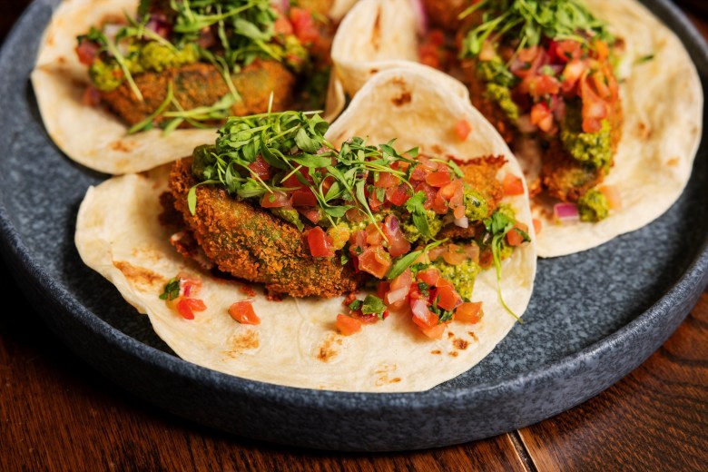 Close-up shot of three tacos topped with pico de gallo and herbs on a gray, speckled ceramic plate.