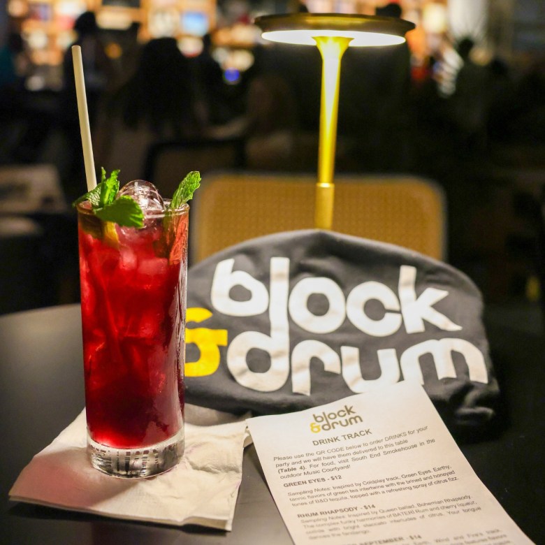 A red colored highball drink garnished with mint leaves sits beside a gold table light atop a paper napkin beside a cocktail menu and a tee shirt reading "block & drum" in Chamblee GA.