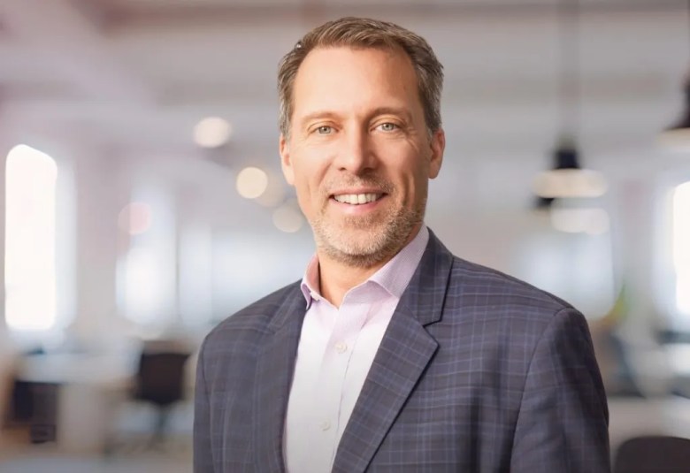Portrait of Doug Shipman wearing a plaid blazer and light shirt, standing in a bright, modern office environment.