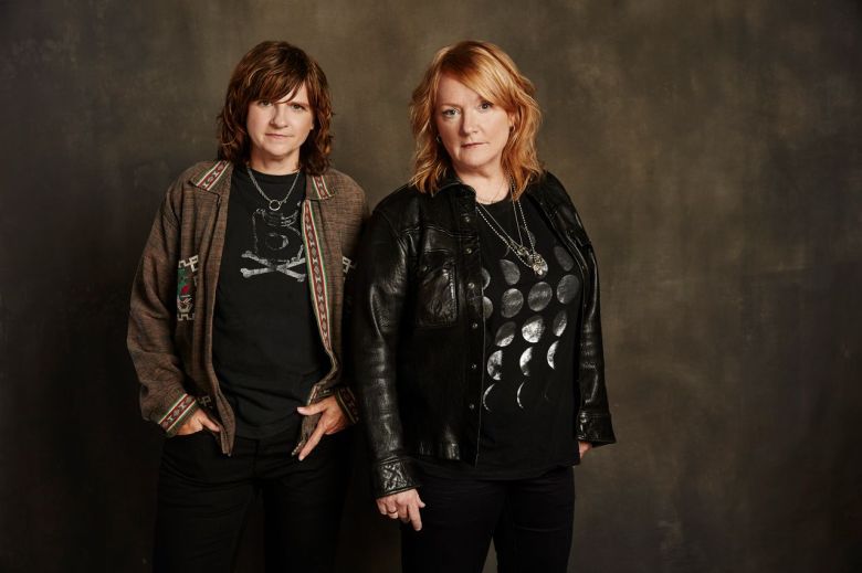 Folk-rock duo Indigo Girls stand looking at the camera against a brown, dappled backdrop.