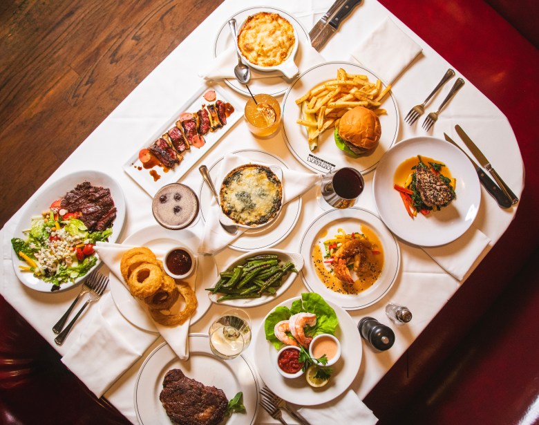 Overhead shot of various steakhouse dishes, including steaks, French onion soup, and shrimp cocktail, over a clothed table in a dining booth. 