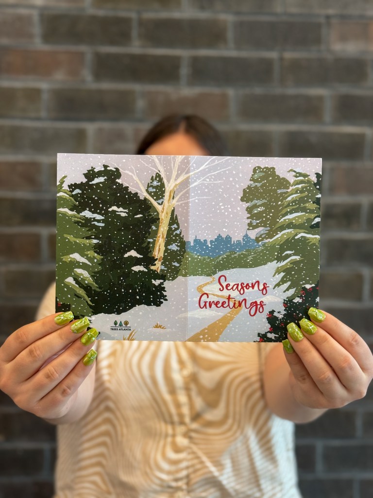 Person holding Trees Atlanta holiday card featuring Elizabeth Lang’s winter landscape with evergreens framing a snowy path toward the Atlanta skyline.