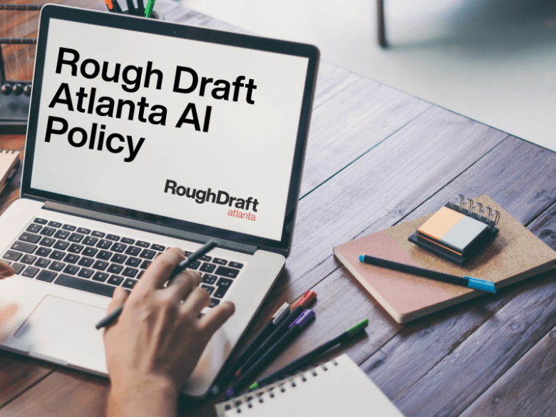 Alt description: A laptop screen displays the text "Rough Draft Atlanta AI Policy" with "RoughDraft atlanta" written below. The laptop is on a wooden desk with various office supplies like pens, notebooks, and a small calendar visible. A hand is seen typing on the laptop keyboard.