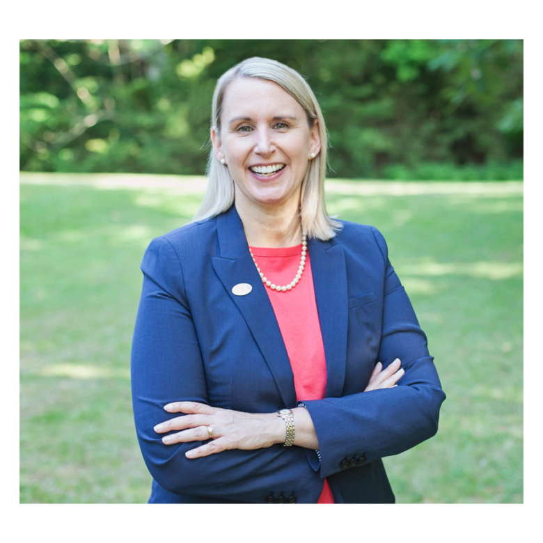 A woman with straight blonde hair smiles while standing outdoors on a lawn with trees in the background. She is wearing a navy blazer, coral top, pearl necklace, and watch, with her arms crossed confidently.