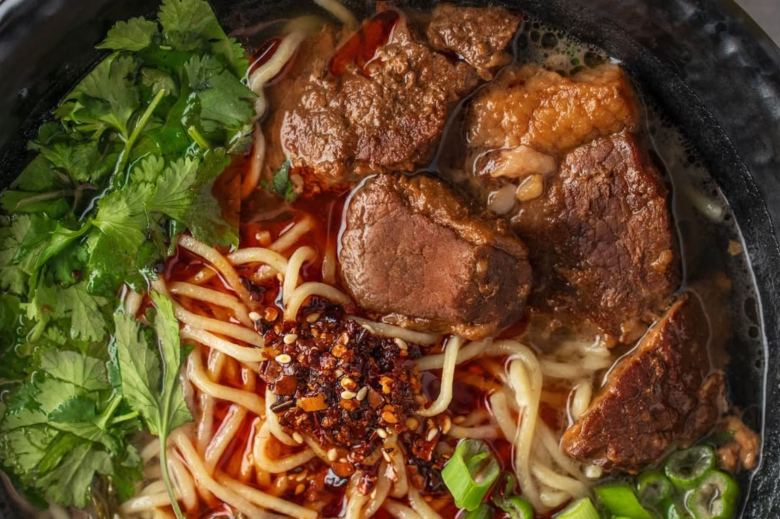 Overhead shot of Szechuan beef noodle soup with chunks of stew meat, noodles, cilantro, chili crisp, and sliced scallions in a deep black bowl. 