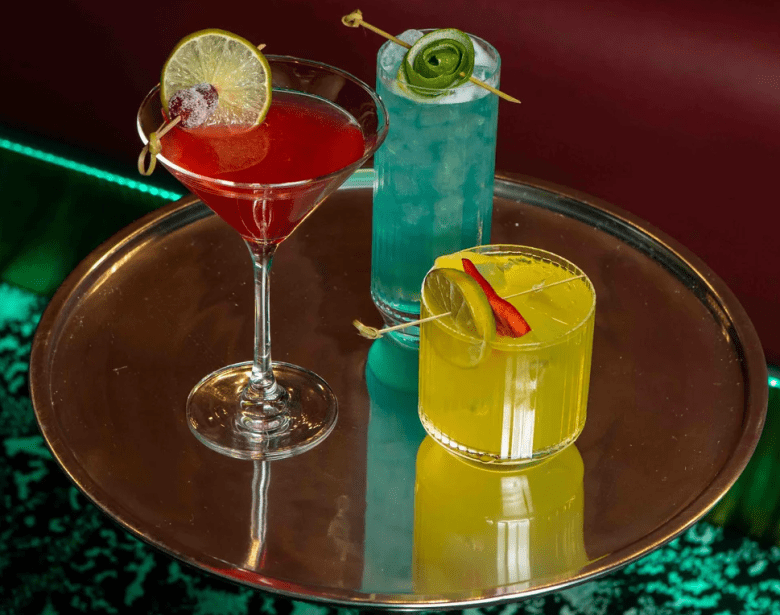 Shot of different cocktails in a martini glass, tall glass, and rocks glass on a mirrored tray. 